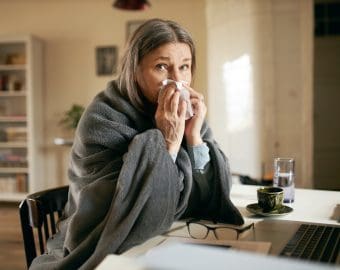 Frustrated gray haired retired woman sitting at table using portable computer to contact physician online, blowing nose, suffering from flu or cold. Technology, health care and elderly people concept