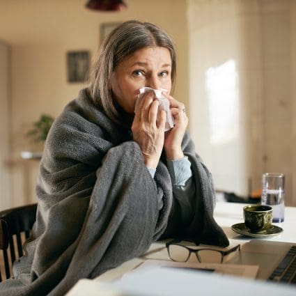 Frustrated gray haired retired woman sitting at table using portable computer to contact physician online, blowing nose, suffering from flu or cold. Technology, health care and elderly people concept